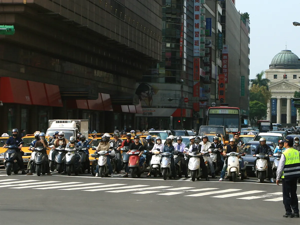 台北館前路口，一整排機車騎士戴著安全帽停在斑馬線前等紅燈，旁邊有交通警察指揮